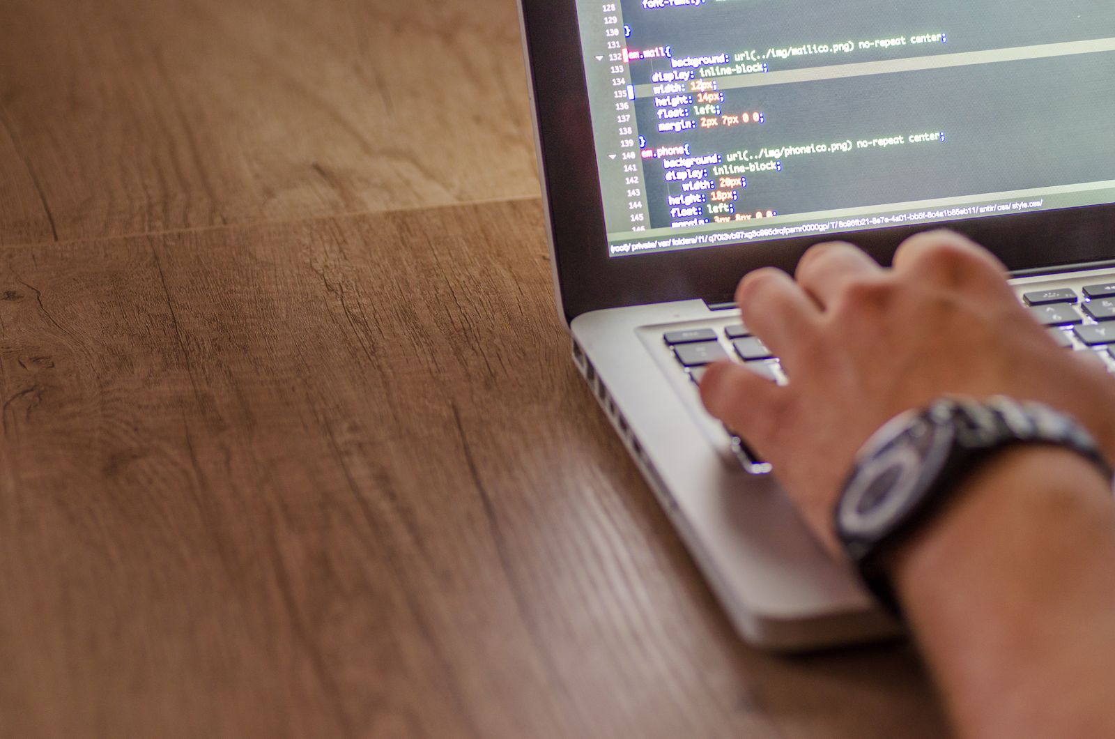 Mão com relógio de pulso digitando em um notebook que mostra linhas de código sobre uma mesa de madeira