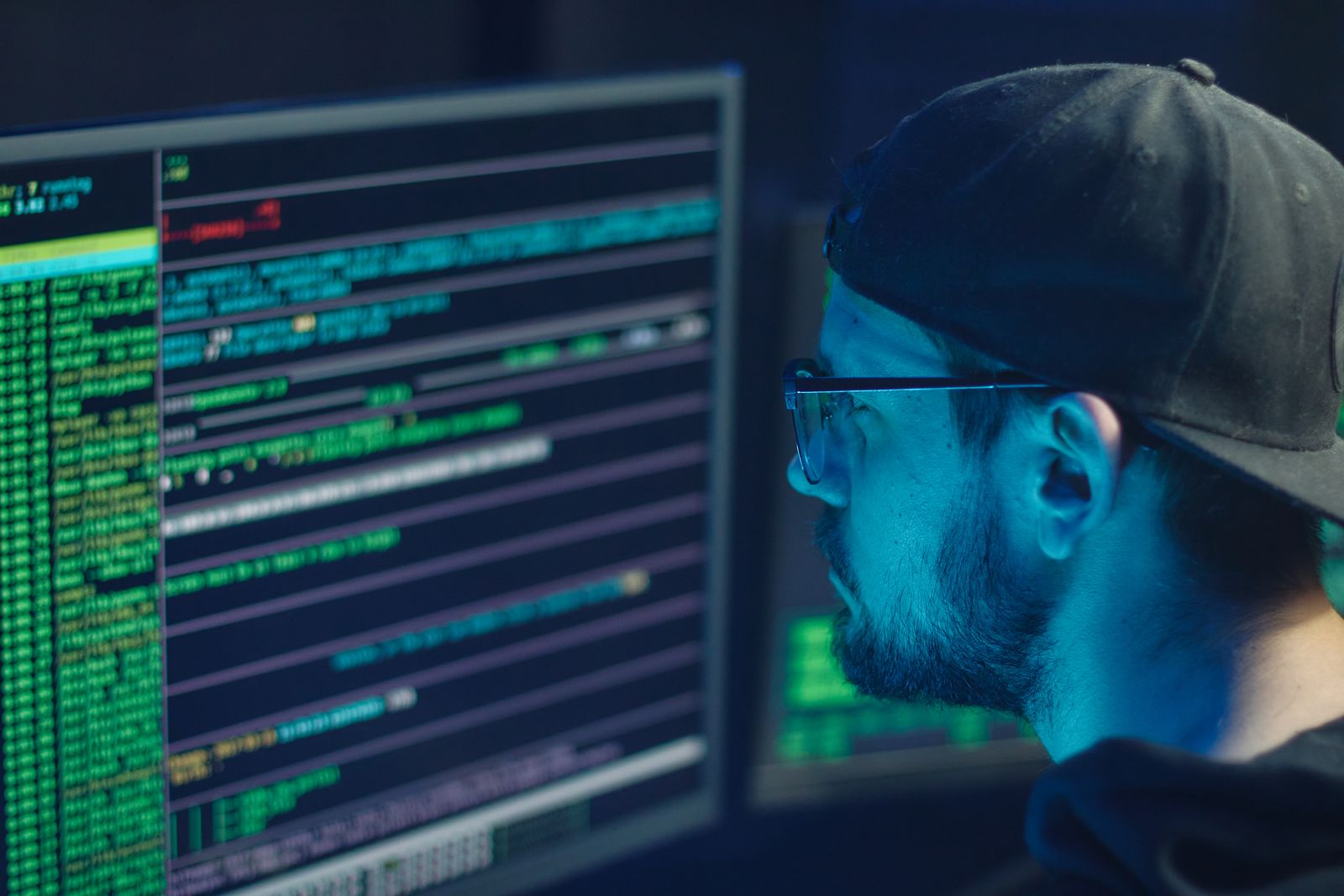 Homem de barba, boné e óculos, de perfil, olhando monitores com texto verde de terminal