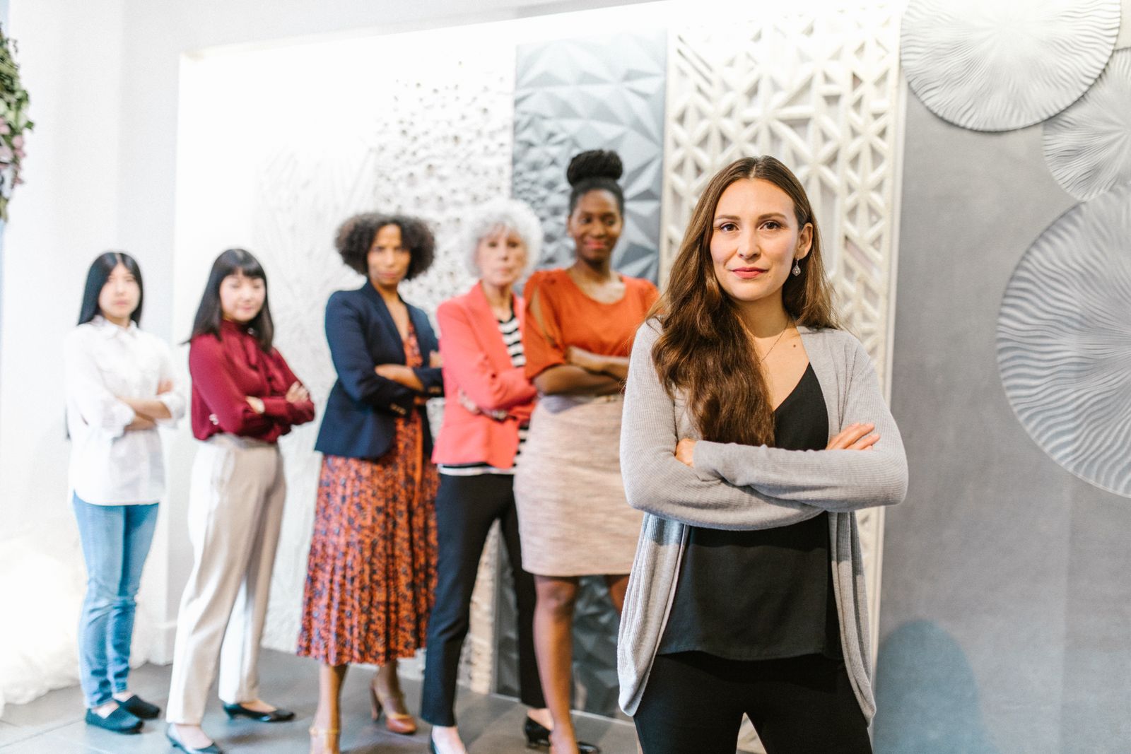 Seis mulheres em pé em uma sala clara, de braços cruzados, uma à frente e cinco atrás