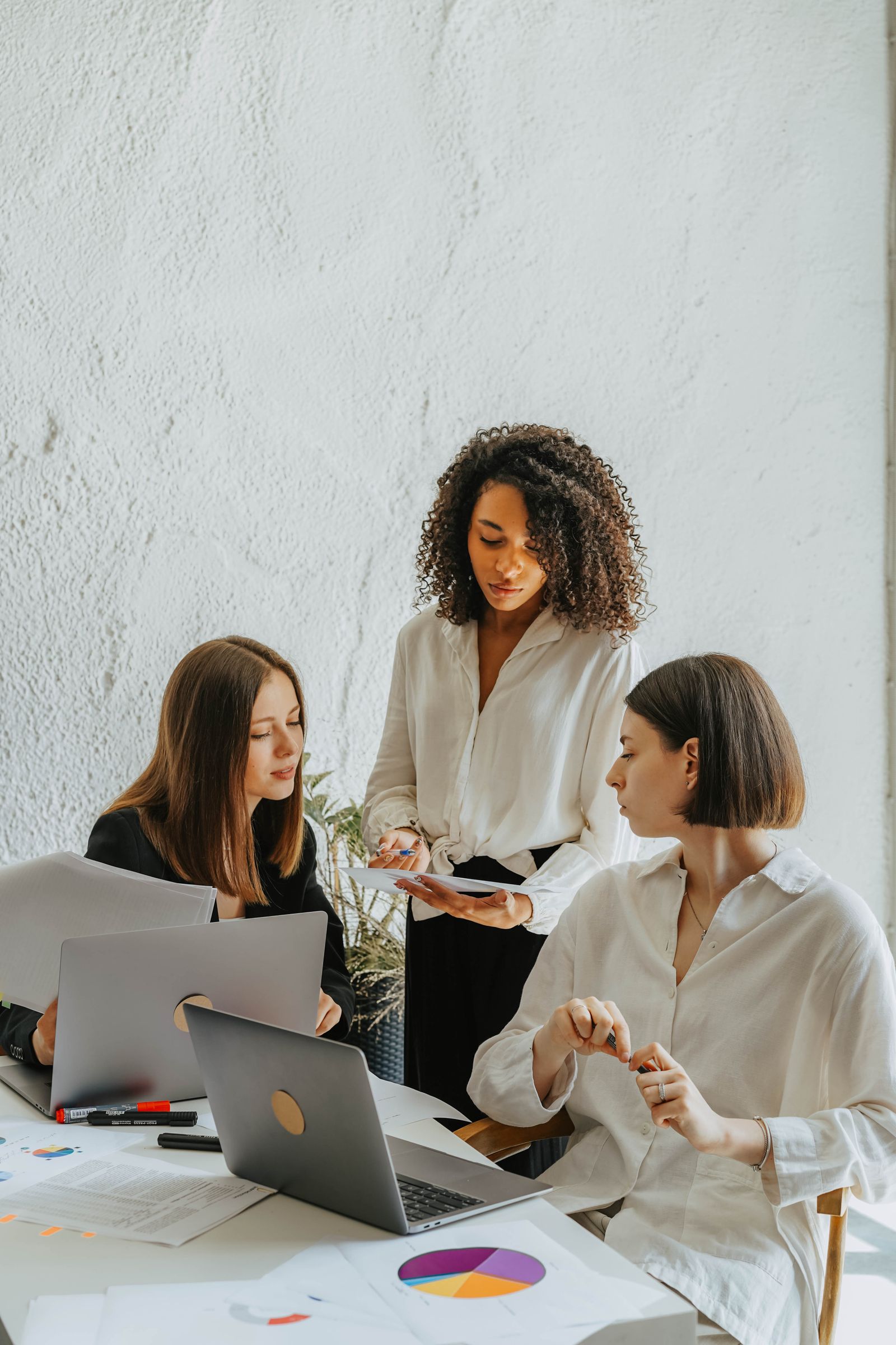 Três mulheres trabalham ao redor de uma mesa com dois notebooks, gráficos impressos e documentos em um escritório claro