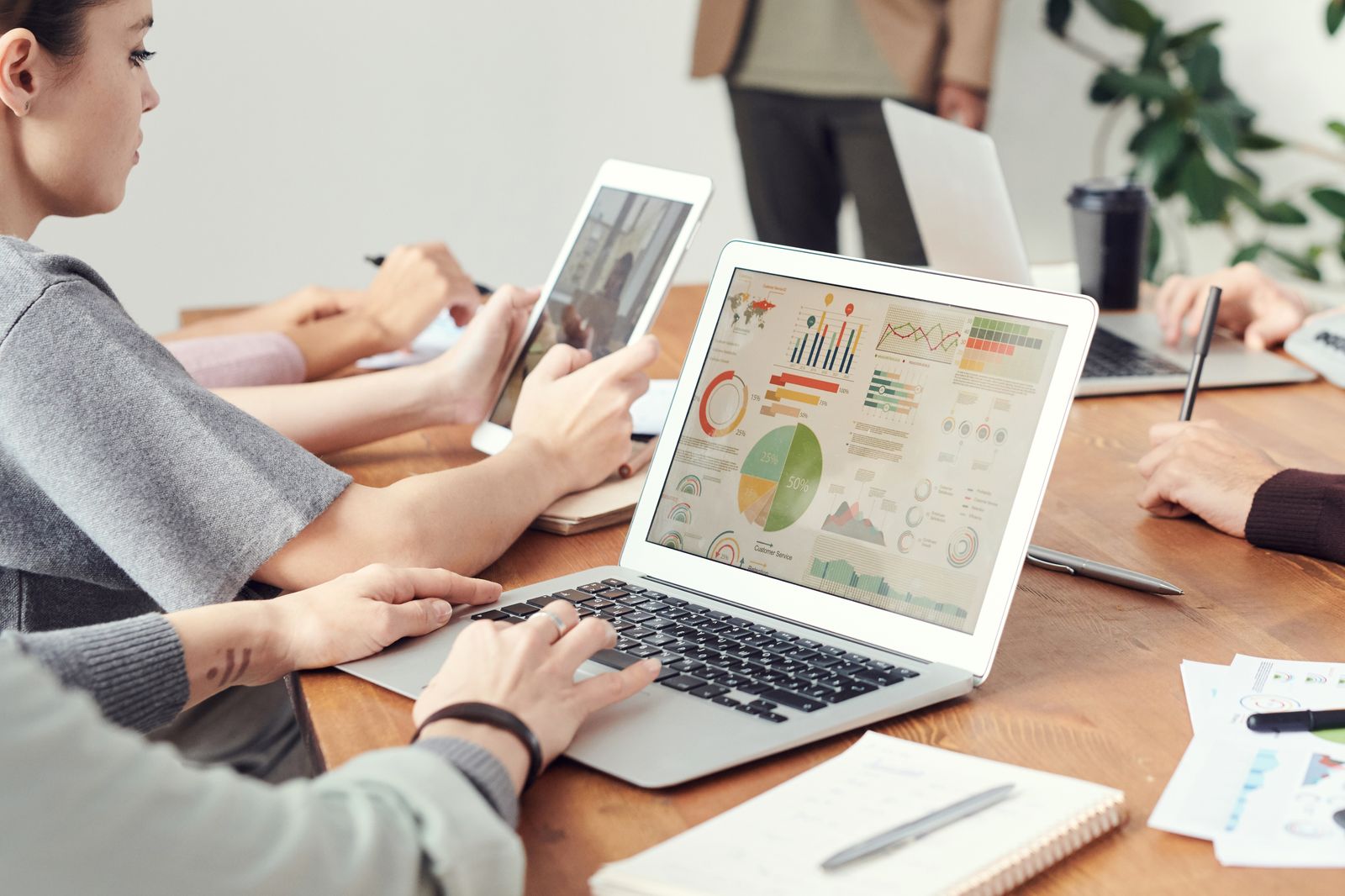 Várias pessoas em uma mesa de madeira usam um notebook com infográficos, um tablet e cadernos