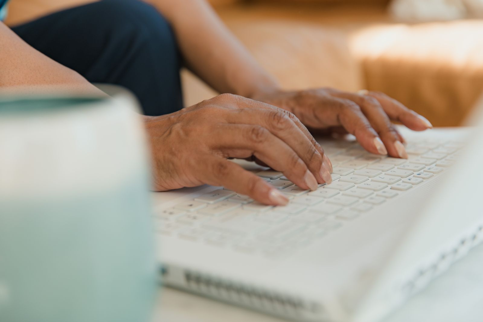 Mãos digitando em um teclado de notebook branco ao lado de uma caneca desfocada